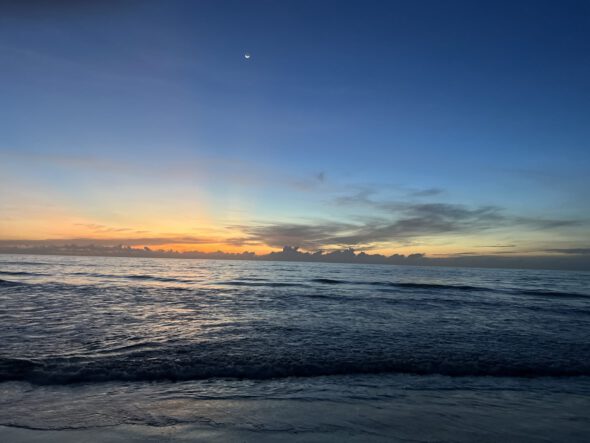 A sunset over the ocean with a crescent moon in the sky.