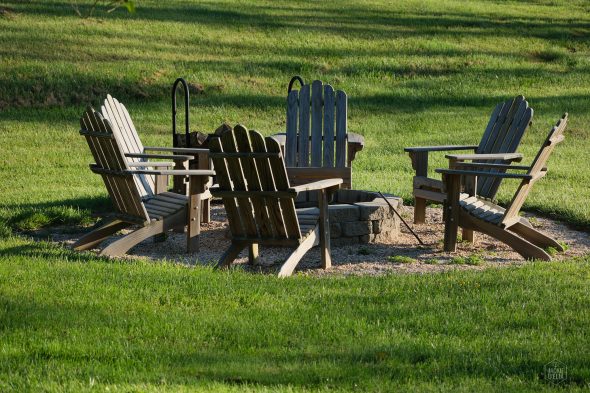 Six wooden Adirondack chairs arranged around a stone fire pit on a neatly mowed grassy area.