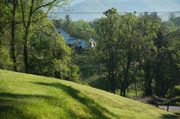A white house is partially visible among lush green trees on a sunny day, with a grassy hill in the foreground and distant mountains in the background.