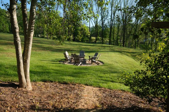 Five Adirondack chairs arranged around a fire pit on a grassy field, surrounded by trees.