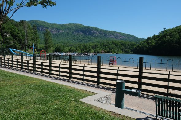 A fenced beach area with a slide and a lifeguard chair, adjacent to a lake, surrounded by green hills and trees under a clear blue sky.
