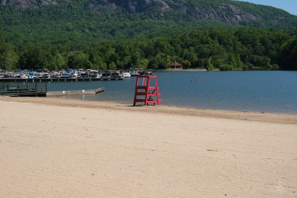 Empty sandy beach with a red lifeguard chair and a docked boat area in the background, surrounded by lush green trees and a rocky hillside.