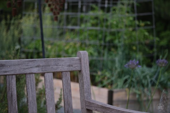 A wooden bench with a weathered finish is positioned in front of a lush garden with various green plants and a trellis structure.