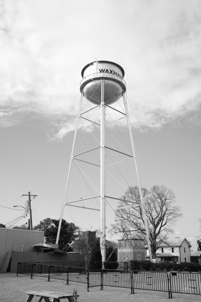 Black and white image of a tall, metal water tower labeled "WAXHAW," with a small town and trees in the background.