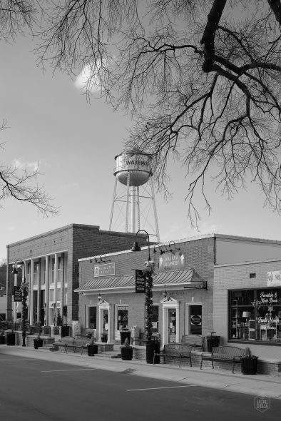 Black and white image of a small town street with a water tower labeled "MATWAN," bare trees, brick buildings, and storefronts.