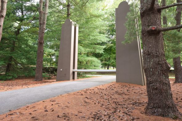 A tall, modern stone gate flanked by trees on a pine-covered road.