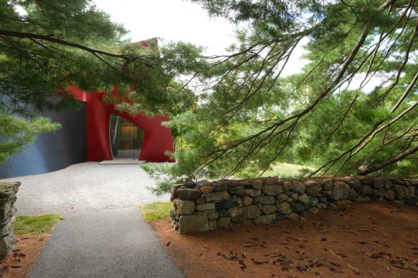 Pathway leading to a modern red and gray building, partially obscured by green pine tree branches, with a stone wall in the foreground.