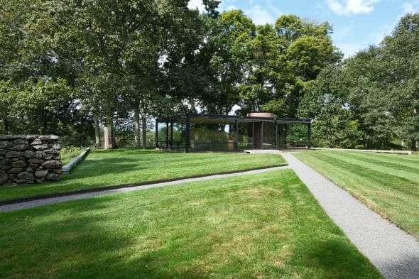 A modern glass house stands surrounded by lush green grass and trees under a blue sky. A stone wall borders the left side. A path leads to the front of the house.