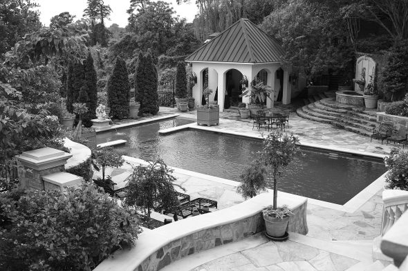 A rectangular swimming pool is surrounded by stone paving, potted plants, and a small pool house with arches and a metal roof, set in a landscaped garden.