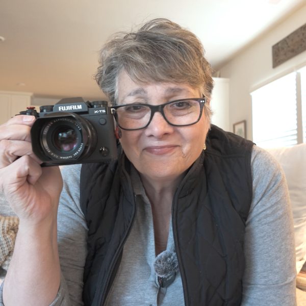 A person with short gray hair and glasses smiles while holding a Fujifilm X-T5 camera up to their face inside a well-lit room.